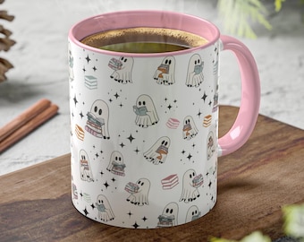 May include: A white ceramic mug with a pink interior and handle. The mug features a repeating pattern of cartoon ghosts holding books, with stars and book illustrations. Steam rises from the mug, suggesting a warm beverage.