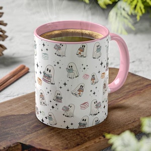 May include: A white ceramic mug with a pink interior and handle. The mug features a repeating pattern of cartoon ghosts holding books, with stars and book illustrations. Steam rises from the mug, suggesting a warm beverage.