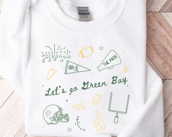 Puede incluir: Sudadera blanca de cuello redondo con gráficos de fútbol en verde y amarillo. El diseño incluye un casco de fútbol, postes de portería y el texto "Let's go Green Bay". La sudadera también tiene el texto "The Pack".