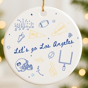 May include: White ceramic ornament with gold string, featuring blue and yellow football-themed illustrations. The text "Let's go Los Angeles" is written in blue script, surrounded by stars and football-related graphics.