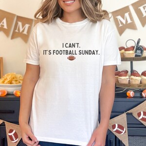 K&ouml;nnte beinhalten: Wei&szlig;es T-Shirt mit dem Text "I can't. It's football Sunday." und einer Football-Grafik. Das Shirt ist Teil einer Football-Party mit Dekorationen wie Bannern und Cupcakes.