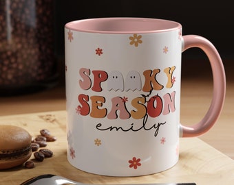 May include: White ceramic mug with a pink interior and handle. The mug features the text "Spooky Season" in retro-style lettering, with ghost and bat graphics. The name "emily" is printed below the text, with floral accents.