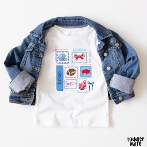 May include: White toddler t-shirt with a blue denim jacket. The shirt features a collection of illustrated stamps with a football helmet, bow, football, buffalo, pennant, and a foam finger. Text on the shirt includes "Buffalo" and "Go Buffalo!"
