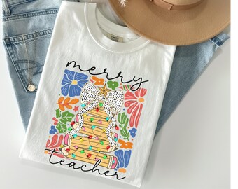 May include: White t-shirt with a colorful graphic design featuring a Christmas tree made of pencils, a polka-dot bow, and the words "merry teacher". The design includes flowers and the shirt is paired with blue jeans and a hat.