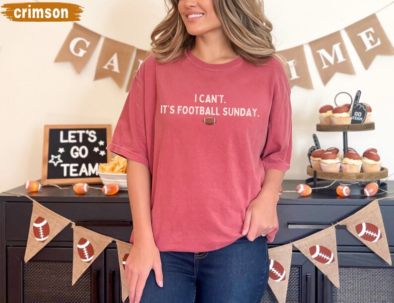 K&ouml;nnte beinhalten: Karmesinrotes T-Shirt mit dem Text "I can't. It's Football Sunday." und einer Football-Grafik. Das Shirt wird mit dunkelblauen Jeans kombiniert. Der Hintergrund zeigt Football-Dekorationen und Banner, ideal f&uuml;r den Spieltag.