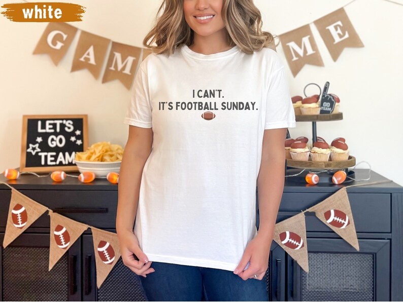 K&ouml;nnte beinhalten: Wei&szlig;es T-Shirt mit dem Text "I can't. It's football Sunday." und einer Football-Grafik. Das Shirt ist Teil einer Football-Party mit Dekorationen wie Bannern und Cupcakes.