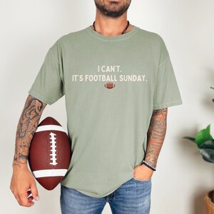 K&ouml;nnte beinhalten: Salbeigr&uuml;nes T-Shirt mit dem Text "I CAN'T. IT'S FOOTBALL SUNDAY." und einer Football-Grafik. Das Shirt wird mit einer blauen Jeans kombiniert. Ein brauner und wei&szlig;er Football wird im Bild gehalten.
