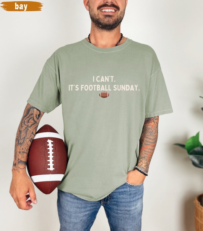 K&ouml;nnte beinhalten: Salbeigr&uuml;nes T-Shirt mit dem Text "I CAN'T. IT'S FOOTBALL SUNDAY." und einer Football-Grafik. Das Shirt wird mit einer blauen Jeans kombiniert. Ein brauner und wei&szlig;er Football wird im Bild gehalten.