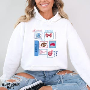 May include: White hoodie featuring a collection of Buffalo-themed graphics. The design includes a football, a helmet, a ribbon, and the word "Buffalo" in blue. The hoodie also has the text "GO BUFFALO!" and a foam finger graphic.