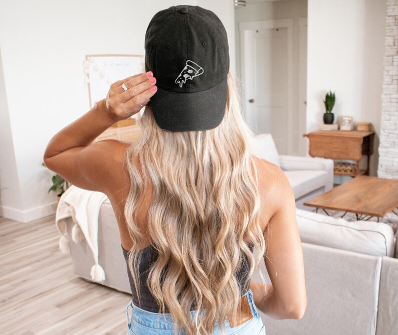 Op de afbeelding: Een zwarte baseballpet met een wit geborduurd pizzapuntontwerp. De pet wordt gedragen door een persoon met lang blond haar.