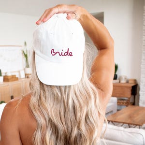 May include: White baseball cap with the word "bride" embroidered in burgundy on the front.