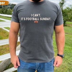 K&ouml;nnte beinhalten: Dunkelgraues T-Shirt mit dem Text "I CAN'T. IT'S FOOTBALL SUNDAY." und einer Football-Grafik. Das Shirt ist einfarbig und hat kurze &Auml;rmel. Die Person tr&auml;gt eine blaue Jeans und eine schwarze Uhr.