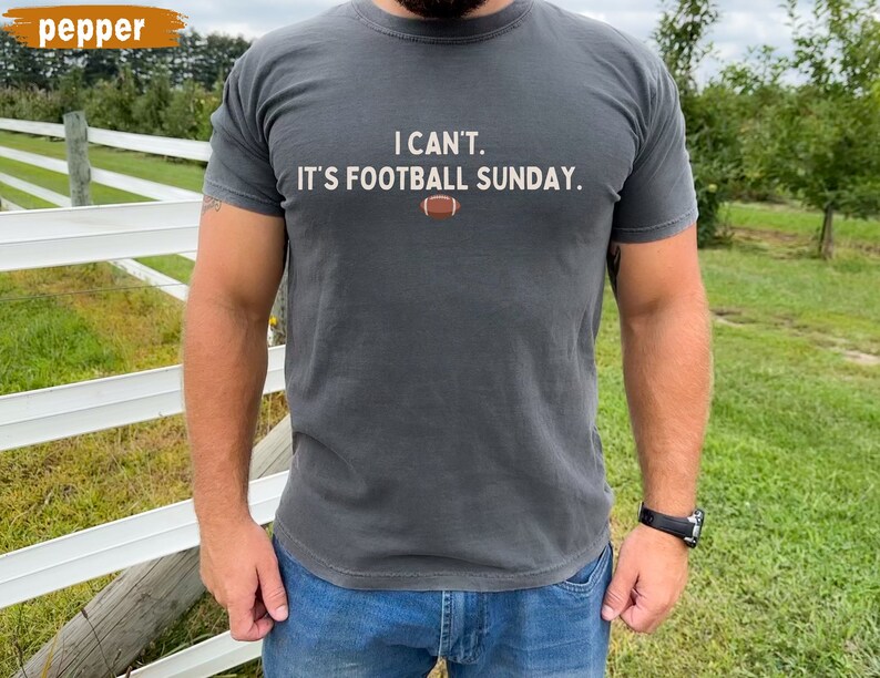 K&ouml;nnte beinhalten: Dunkelgraues T-Shirt mit dem Text "I CAN'T. IT'S FOOTBALL SUNDAY." und einer Football-Grafik. Das Shirt ist einfarbig und hat kurze &Auml;rmel. Die Person tr&auml;gt eine blaue Jeans und eine schwarze Uhr.