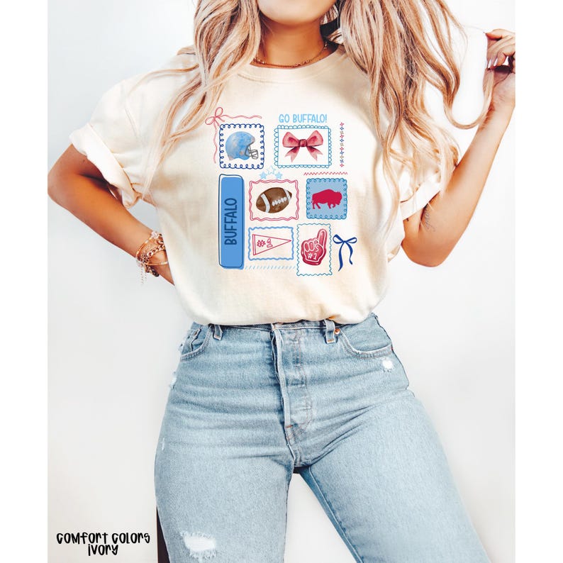 May include: Ivory-colored t-shirt with a graphic design featuring various Buffalo-themed illustrations. The design includes a football, a helmet, a bow, and the word "Buffalo" in a blue rectangle. The text "GO BUFFALO!" is also present.