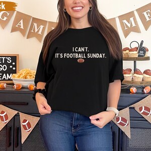 K&ouml;nnte beinhalten: Schwarzes T-Shirt mit dem Text "I can't. It's football Sunday." und einer Football-Grafik. Das Bild enth&auml;lt dekorative Banner mit Footballs und den W&ouml;rtern "GAME" und "ME".