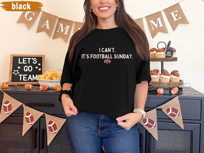 K&ouml;nnte beinhalten: Schwarzes T-Shirt mit dem Text "I can't. It's football Sunday." und einer Football-Grafik. Das Bild enth&auml;lt dekorative Banner mit Footballs und den W&ouml;rtern "GAME" und "ME".