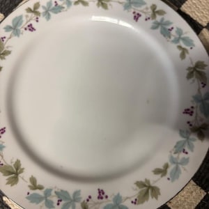 Puede incluir: Un plato de cena de cerámica blanca con un borde plateado y un delicado diseño floral con hojas verdes y bayas moradas.