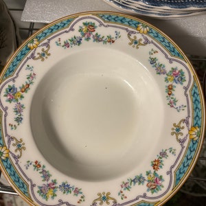 Pode incluir: Uma tigela de sopa de porcelana branca com um padrão floral azul e amarelo ao redor da borda. A tigela tem um interior branco e uma borda ligeiramente elevada.
