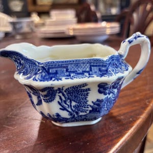 Op de afbeelding: Een blauw-witte keramische creamer met een handvat en tuit. De creamer heeft een gedetailleerd Willow-patroon. De creamer staat op een houten oppervlak.