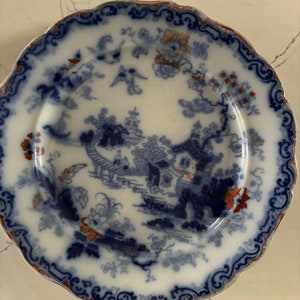 Puede incluir: Un plato de cerámica blanco con un diseño floral azul y rojo. El plato presenta una imagen central de una pagoda azul y blanca con un puente y árboles.