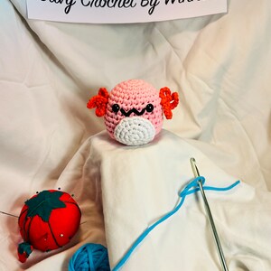 May include: A pink and white crocheted axolotl plushie with orange gills. The plushie is sitting on a white fabric surface with a blue yarn ball, a red pincushion, and a crochet hook with blue yarn.