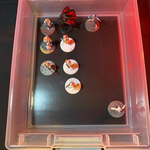May include: A clear plastic storage bin containing several miniature figurines. The figurines depict various characters, some with red and black wings, others in blue and white armor, and others in orange and white uniforms. The figurines are on circular bases.