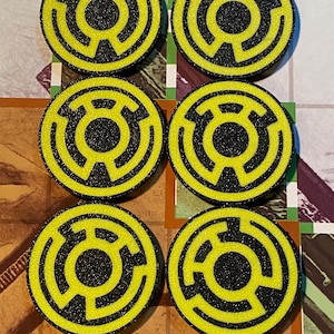 Op de afbeelding: Zes gele en zwarte ronde spelstukken met een doolhofachtig ontwerp. De stukken zijn gerangschikt in twee rijen van drie. Het ontwerp heeft een zwarte cirkel in het midden, omgeven door een gele doolhof.