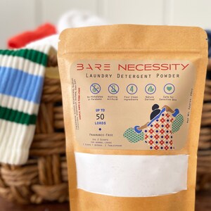 Op de afbeelding: Een bruine papieren zak met een wit label waarop "BARE NECESSITY LAUNDRY DETERGENT POWDER" staat. Het label heeft pictogrammen voor geen ftalaten of parabenen, niets kunstmatigs, vier schone ingredi&euml;nten, van nature afkomstig en veilig voor gevoelige huid. De zak heeft een witte schep met een houten handvat en een hoop wit poeder ervoor.
