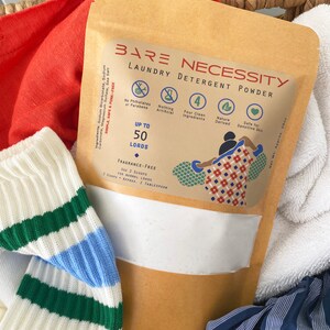 Op de afbeelding: Een bruine papieren zak met een wit label waarop "Bare Necessity Laundry Detergent Powder" staat. Het label somt de volgende kenmerken op: "Tot 50 wasbeurten", "Geurvrij", "Geen ftalaten of parabenen", "Niets kunstmatigs", "Vier schone ingredi&euml;nten", "Natuurlijk afgeleid" en "Veilig voor gevoelige huid".