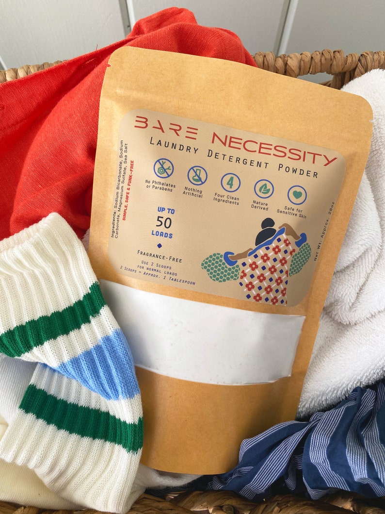 Op de afbeelding: Een bruine papieren zak met een wit label waarop "Bare Necessity Laundry Detergent Powder" staat. Het label somt de volgende kenmerken op: "Tot 50 wasbeurten", "Geurvrij", "Geen ftalaten of parabenen", "Niets kunstmatigs", "Vier schone ingredi&euml;nten", "Natuurlijk afgeleid" en "Veilig voor gevoelige huid".