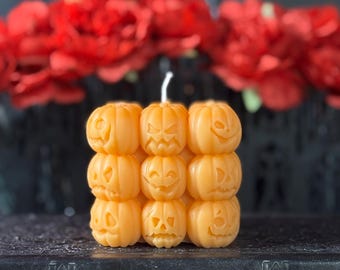 Puede incluir: Una vela decorativa con forma de nueve calabazas naranjas con caras talladas, apiladas en un cubo. La vela tiene una mecha blanca y está colocada sobre una superficie oscura, con flores rojas borrosas en el fondo. Decoración de Halloween.