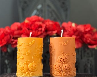 Puede incluir: Dos velas de pilar naranjas con tallas temáticas de Halloween. Una vela es de un tono naranja más claro y presenta murciélagos y calabazas. La otra vela es de un tono naranja más oscuro y tiene calabazas talladas. Ambas velas tienen mechas blancas.