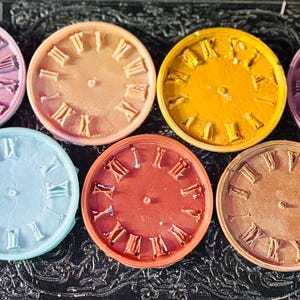 May include: A collection of seven colorful clock faces, each with Roman numerals marking the hours. The faces are in shades of pink, orange, yellow, purple, blue, red, and brown. The clock faces are arranged in a circular pattern.
