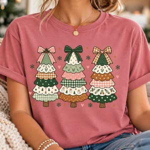 May include: A dusty rose short-sleeved t-shirt featuring a graphic of three stylised Christmas trees. Each tree is decorated with a large bow at the top and layered with various patterns including gingham, floral, and solid colours with small icons.
