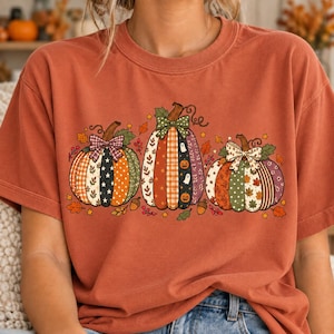 May include: A rust-coloured t-shirt featuring a graphic print of three pumpkins. Each pumpkin is decorated with a different pattern, including plaid, polka dots, and stars, and is topped with a bow. Small autumn leaves and acorns surround the pumpkins.