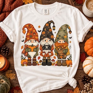 Op de afbeelding: Een wit t-shirt met korte mouwen en een afbeelding van drie kabouters met lange witte baarden. De linker kabouter draagt een oranje muts met bladeren, de middelste een zwarte muts met pompoenen en de rechter een groene muts met spinnenwebben.