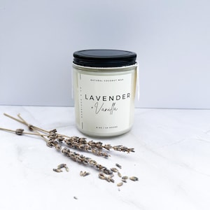 Puede incluir: Una vela blanca en un tarro de vidrio transparente con tapa negra. La vela está etiquetada con "Lavender + Vanilla" y "Natural Coconut Wax". La vela es de 8 oz y tiene un tiempo de combustión de 40 horas. Flores de lavanda secas están esparcidas sobre una superficie blanca.