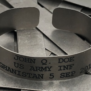 May include: A silver-colored cuff bracelet with engraved text. The text includes the name "JOHN Q. DOE", "US ARMY INF", "AFGHANISTAN 5 SEP 2015", and a small emblem. The bracelet is open-ended and sits on a surface of similar metal strips.