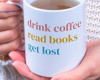 May include: White ceramic mug with the text "drink coffee read books get lost" in colorful lettering. The mug is filled with a light brown liquid.