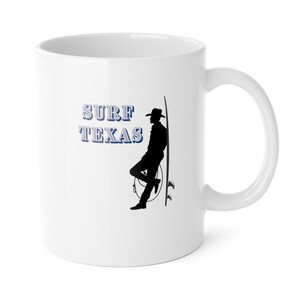 May include: White ceramic mug with a black silhouette of a person leaning against a surfboard. The text "SURF TEXAS" is printed in blue and white. The mug has a curved handle.