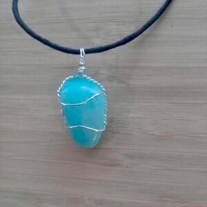 May include: A turquoise gemstone pendant wrapped in silver wire, hanging from a black cord necklace.
