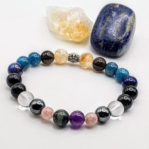 May include: A beaded bracelet featuring a variety of colorful gemstones, including blue lapis lazuli, clear quartz, and amethyst. The bracelet is strung on an elastic cord and has a silver-colored spacer bead. A yellow-orange crystal and a blue stone are also present.