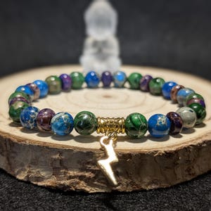May include: A multi-colored beaded bracelet with a gold lightning bolt charm. The beads are green, blue, and purple with a marbled pattern. The bracelet is on a wooden surface.