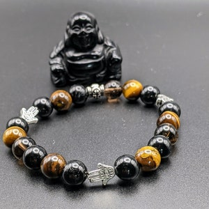 May include: A black and gold beaded bracelet with silver hamsa hand charms. The bracelet is made with tiger's eye and black onyx beads.