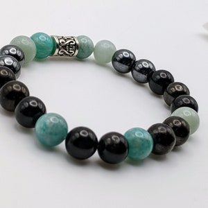 May include: A beaded bracelet with black, green, and gray stones. The bracelet has a silver charm in the center.