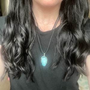 May include: A turquoise gemstone pendant necklace with a silver wire wrap. The necklace is on a black cord and is being worn by a person with long black hair.