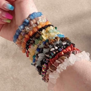 May include: A stack of colorful gemstone bracelets, including blue, green, orange, red, and white stones. The bracelets are made of various materials, including beads, chips, and smooth stones.