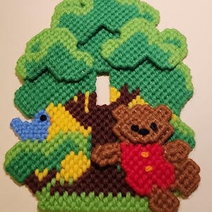 Puede incluir: Un oso de peluche marrón que lleva una camisa roja con botones amarillos se sienta frente a un árbol verde con un pájaro azul posado en una rama. El árbol está hecho de hilo y es una decoración de pared.