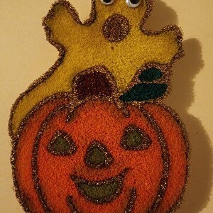 May include: A yellow felt ghost with googly eyes sits on top of an orange felt pumpkin with a carved jack-o'-lantern face. Both are decorated with gold glitter.