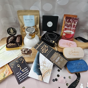 May include: A selection of Ramadan gifts, featuring chocolate-covered dates, Bombay Mix, honey, a black box, prayer beads, tea, cookies, and decorative cards. The items are presented on a wooden board with a ribbon and confetti.